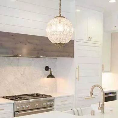 A modern kitchen with a clean, contemporary design. It features stainless steel appliances, white cabinetry with gold handles, a marble backsplash, and a kitchen island with a sink and gold faucet. Spherical pendant lights and a wall-mounted sconce provide stylish lighting.